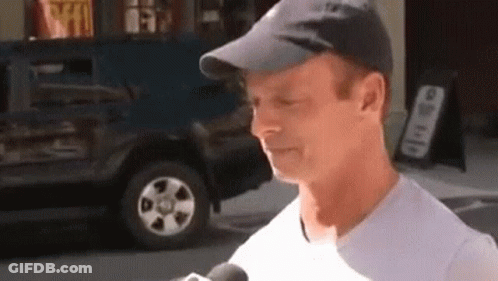 A Man Wearing A Hat And A White Shirt Is Talking Into A Microphone On The Street . GIF