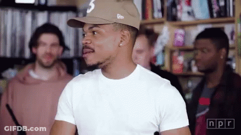 A Man Wearing A Hat And A White T-Shirt Is Standing In A Room With Other People . GIF