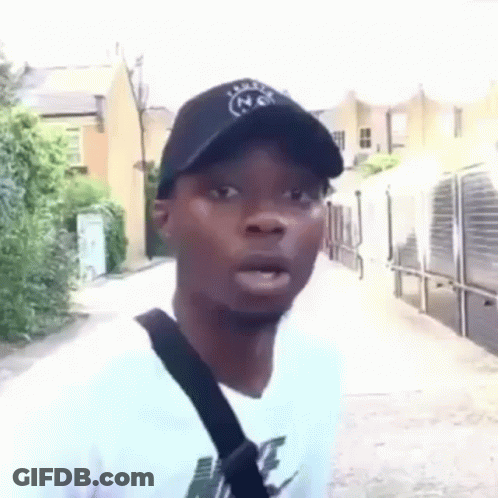 A Man Wearing A Hat And A White T-Shirt Is Standing On A Sidewalk . GIF