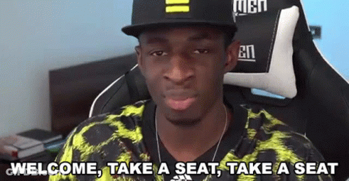 A Man Wearing A Hat And A Yellow Shirt Is Sitting In A Chair And Making A Funny Face . GIF