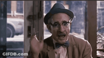A Man Wearing A Hat And Bow Tie Is Sitting In Front Of A Window And Making A Funny Face . GIF