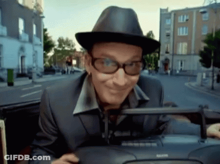 A Man Wearing A Hat And Glasses Is Driving Down A Street GIF