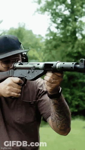 A Man Wearing A Hat And Glasses Is Holding A Gun GIF