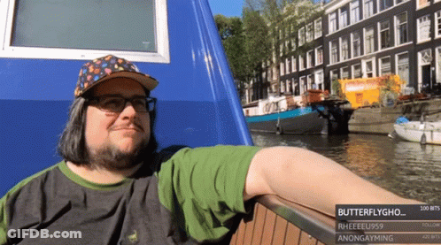 A Man Wearing A Hat And Glasses Is Leaning On A Railing Next To A Body Of Water With Buildings In The Background GIF