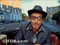 A Man Wearing A Hat And Glasses Is Sitting In Front Of A City Street GIF