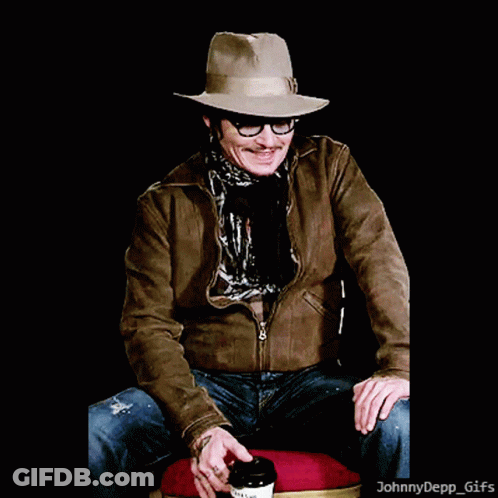 A Man Wearing A Hat And Glasses Is Sitting On A Chair With A Cup Of Coffee GIF