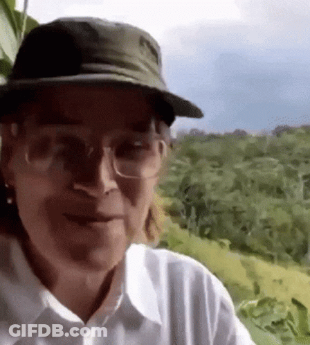 A Man Wearing A Hat And Glasses Is Standing In A Field . GIF