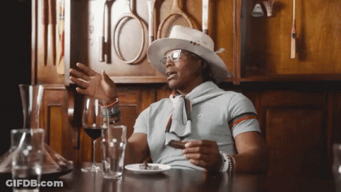 A Man Wearing A Hat And Glasses Sits At A Table With Wine Glasses And A Plate Of Food GIF