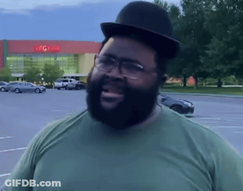 A Man Wearing A Hat And Glasses Stands In A Parking Lot In Front Of A Building That Says The G Rite GIF