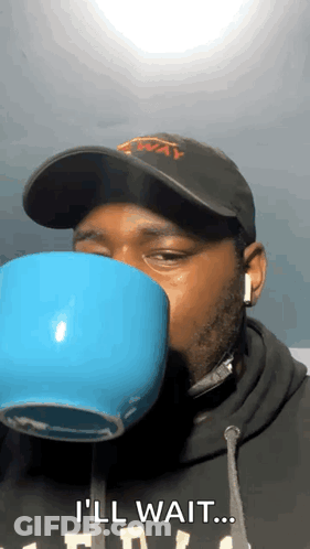 A Man Wearing A Hat And Headphones Is Drinking From A Blue Cup And Saying I 'Ll Wait . GIF
