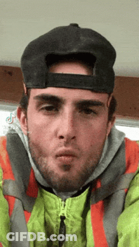 A Man Wearing A Hat And Safety Vest Is Blowing A Kiss . GIF