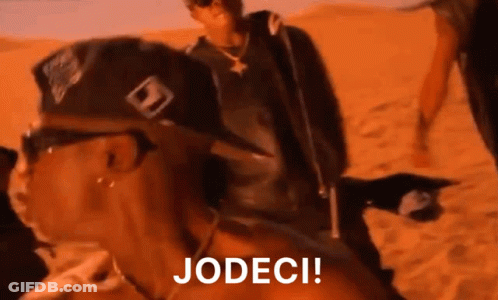 A Man Wearing A Hat And Sunglasses Is Riding A Camel In The Desert While Another Man Looks On . GIF