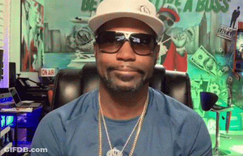 A Man Wearing A Hat And Sunglasses Is Sitting In Front Of A Sign That Says " On Air " GIF
