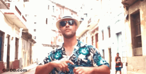 A Man Wearing A Hat And Sunglasses Is Standing In A Street GIF