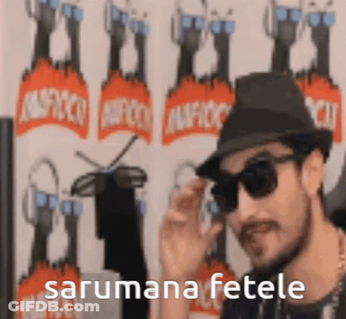 A Man Wearing A Hat And Sunglasses Is Standing In Front Of A Wall With The Words Sarumana Fetele Written On It . GIF