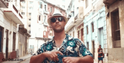 A Man Wearing A Hat And Sunglasses Is Standing On A City Street GIF