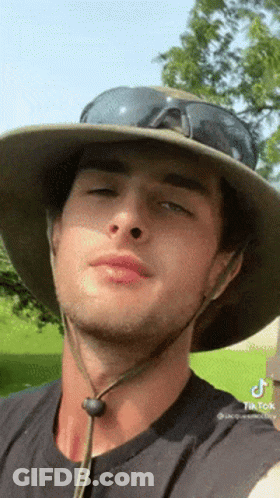 A Man Wearing A Hat And Sunglasses Looks At The Camera . GIF