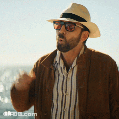 A Man Wearing A Hat And Sunglasses Stands In Front Of A Body Of Water GIF