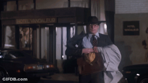 A Man Wearing A Hat And Tie Is Holding A Briefcase With The Letter S On The Front GIF