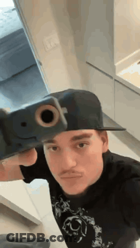 A Man Wearing A Hat Is Holding A Gun To His Face . GIF