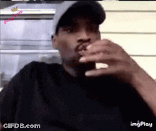 A Man Wearing A Hat Is Smoking A Cigarette While Looking At His Phone . GIF