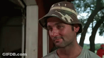 A Man Wearing A Hat Is Standing In Front Of A Building . GIF