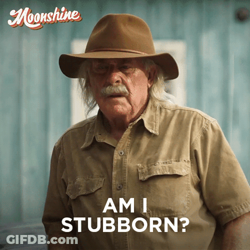 A Man Wearing A Hat Says " Am I Stubborn " In Front Of A Blue Wall GIF