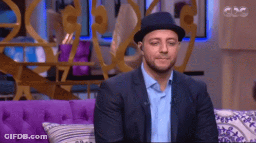 A Man Wearing A Hat Sits On A Purple Couch In Front Of A Cdg Sign GIF