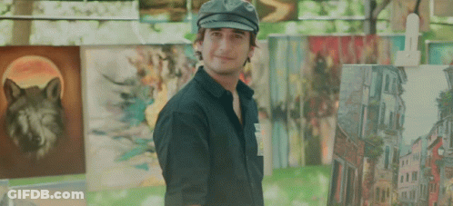 A Man Wearing A Hat Stands In Front Of A Wall Of Paintings Including One Of A Wolf GIF