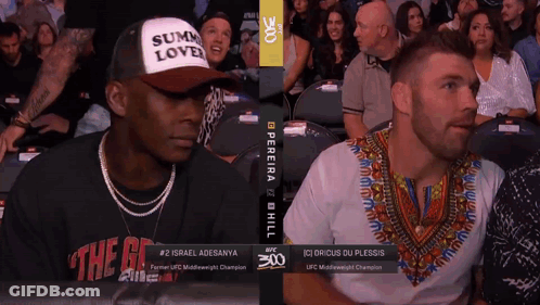 A Man Wearing A Hat That Says Sum Love Sits Next To A Man Wearing A Shirt That Says The G GIF