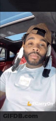 A Man Wearing A Hat With Feathers On It Is Sitting In A Car With #Carryon Written On The Bottom GIF