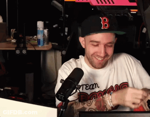 A Man Wearing A Hat With The Letter B On It Is Sitting In Front Of A Microphone And Smiling . GIF