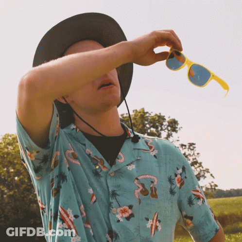A Man Wearing A Hawaiian Shirt And A Hat Holds A Pair Of Yellow Sunglasses GIF