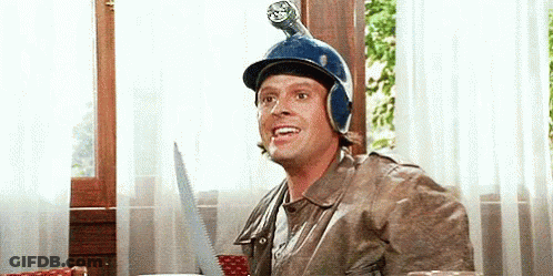 A Man Wearing A Helmet And A Jacket Is Sitting At A Table With A Saw On His Head . GIF