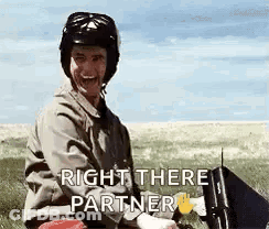 A Man Wearing A Helmet Is Sitting In A Field With A Laptop And A Sign That Says Right There Partner . GIF