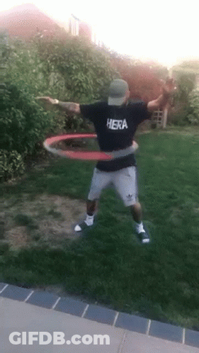 A Man Wearing A Hera Shirt Is Playing With A Hula Hoop In A Yard . GIF