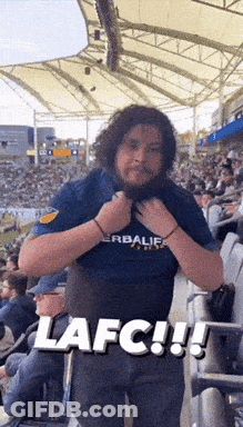 A Man Wearing A Herbalife Shirt Is Standing In A Crowded Stadium GIF
