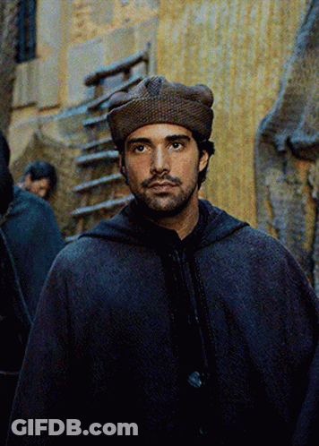 A Man Wearing A Hooded Cape And A Hat Looks At The Camera GIF