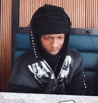 A Man Wearing A Hooded Jacket And A Beanie Is Sitting At A Table . GIF