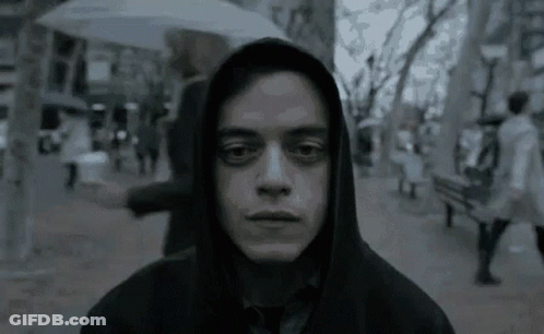 A Man Wearing A Hooded Jacket And Tie Is Standing On A Sidewalk In The Rain . GIF