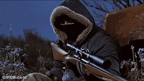 A Man Wearing A Hooded Jacket Is Holding A Sniper Rifle GIF