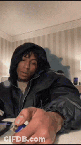 A Man Wearing A Hooded Jacket Is Sitting At A Table Holding A Lighter . GIF