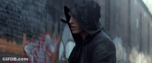 A Man Wearing A Hooded Jacket Is Standing In Front Of A Brick Wall With Graffiti On It GIF