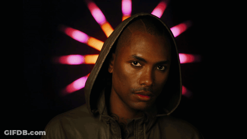 A Man Wearing A Hooded Jacket Stands In Front Of A Pink And Orange Light GIF