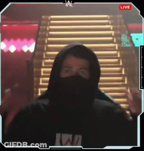 A Man Wearing A Hoodie And A Mask Is Standing In Front Of Stairs And Giving A Peace Sign . GIF