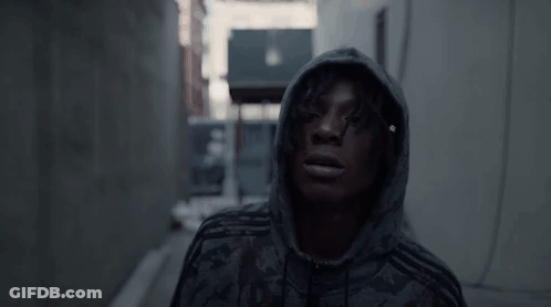 A Man Wearing A Hoodie Is Standing In A Dark Alleyway . GIF