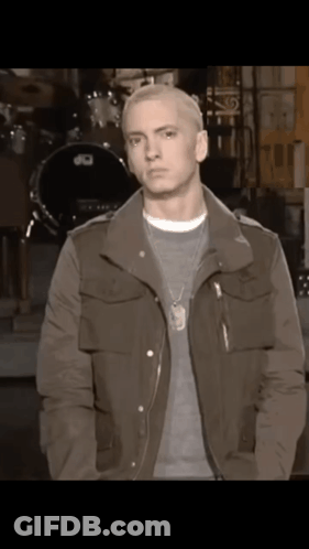 A Man Wearing A Jacket And A Necklace Stands In Front Of A Drum Set .. GIF