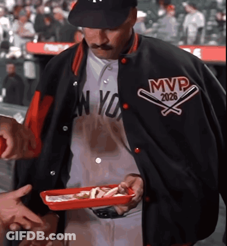 A Man Wearing A Jacket That Says Mvp 2026 Holds A Tray Of Food GIF