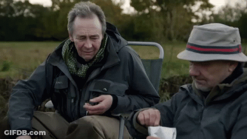 A Man Wearing A Jacket That Says ' Salomon ' On It Sits Next To Another Man Wearing A Hat GIF