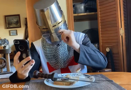 A Man Wearing A Knight 'S Helmet Is Sitting At A Table Eating Bread And Looking At His Phone GIF